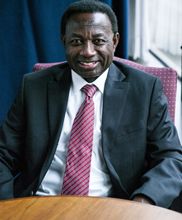 Image of John Abrefah wearing a blue suit jacket with white shirt and red tie