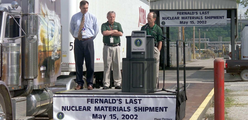 Image of three men with one speaking at podium above banner "Fernald's Last Nuclear Materials Shipment May 15, 2002".